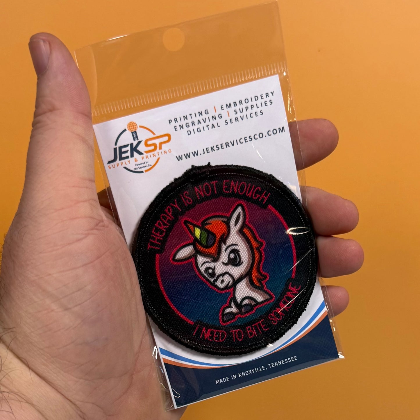A hand holds a packaged embroidered patch against a solid orange background. The patch features a cartoon unicorn with a green horn and pink tongue on a dark blue and black background, with the text 'THERAPY IS NOT ENOUGH' and 'I NEED TO BITE BACK.