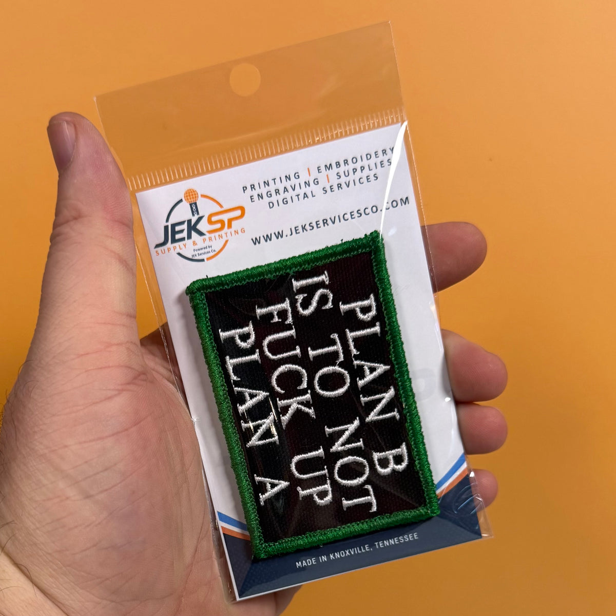 A hand holds a packaged embroidered patch against a solid orange background. The patch reads 'PLAN B IS TO NOT FUCK UP' in white lettering on a black background with a green border.