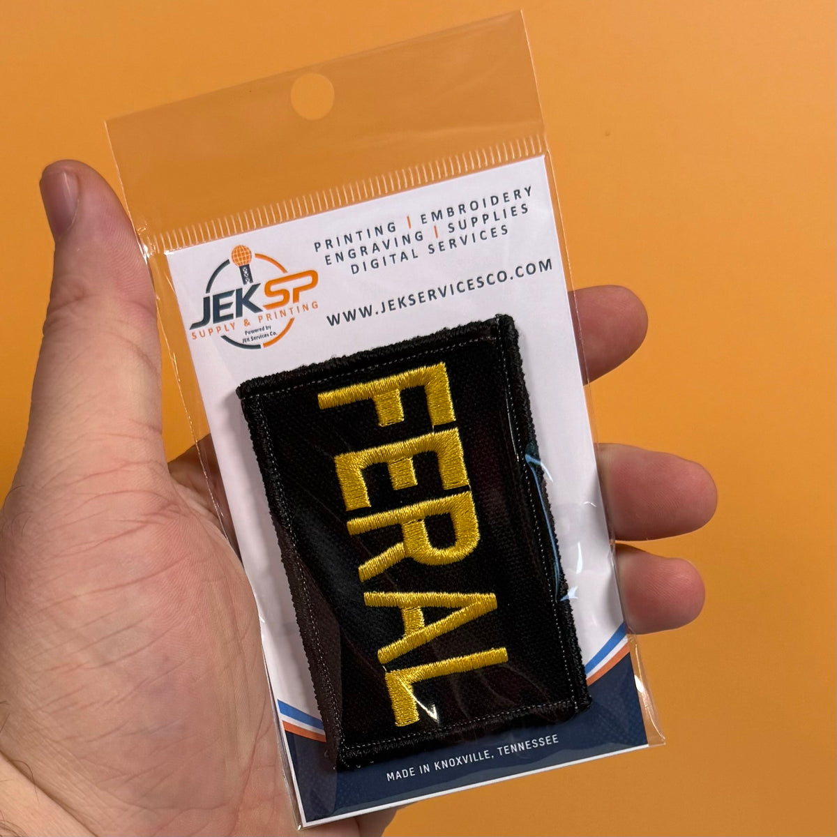 A hand holds a packaged embroidered patch that reads 'FERAL' in yellow on a black background, attached to a clear plastic sleeve with a branded card backing. The card shows the JEKSP brand and text for printing, embroidery, and supplies.