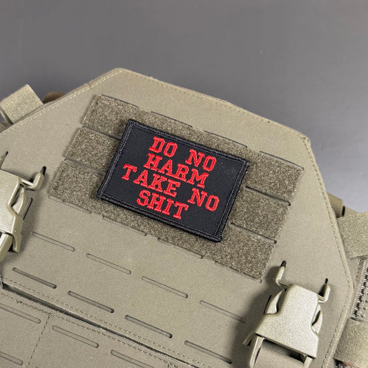 Do No Harm Take No Shit embroidered morale patch: black tactical patch with red embroidered text reading 'DO NO HARM TAKE NO SHIT' attached to a tan webbed vest or plate carrier with plastic quick-release buckles.