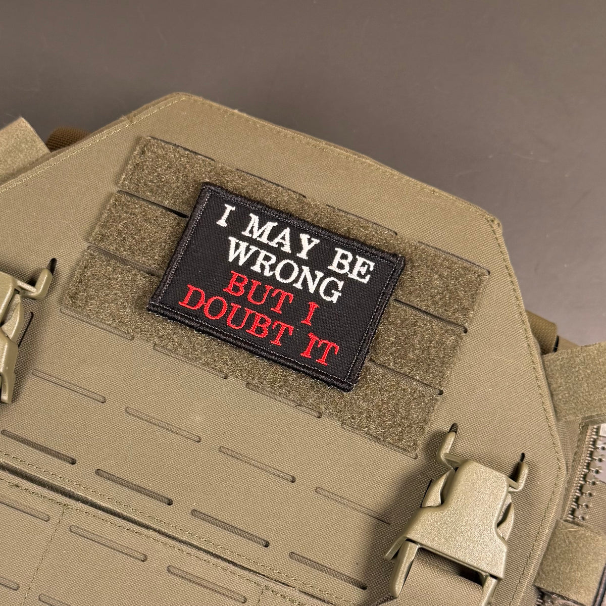 A rectangular black tactical patch with the embroidered text 'I MAY BE WRONG BUT I DOUBT IT' in white and red, shown attached to tan webbing on a tan tactical vest or plate carrier.