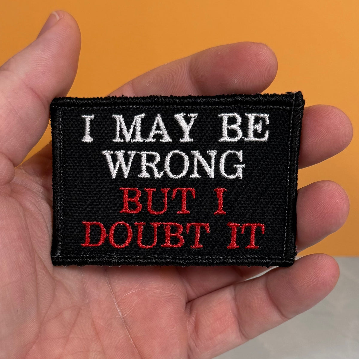 A rectangular black embroidered patch held in a hand, featuring the text 'I MAY BE WRONG BUT I DOUBT IT' in white and red lettering, against an orange background.