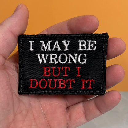 A rectangular black embroidered patch held in a hand, featuring the text 'I MAY BE WRONG BUT I DOUBT IT' in white and red lettering, against an orange background.