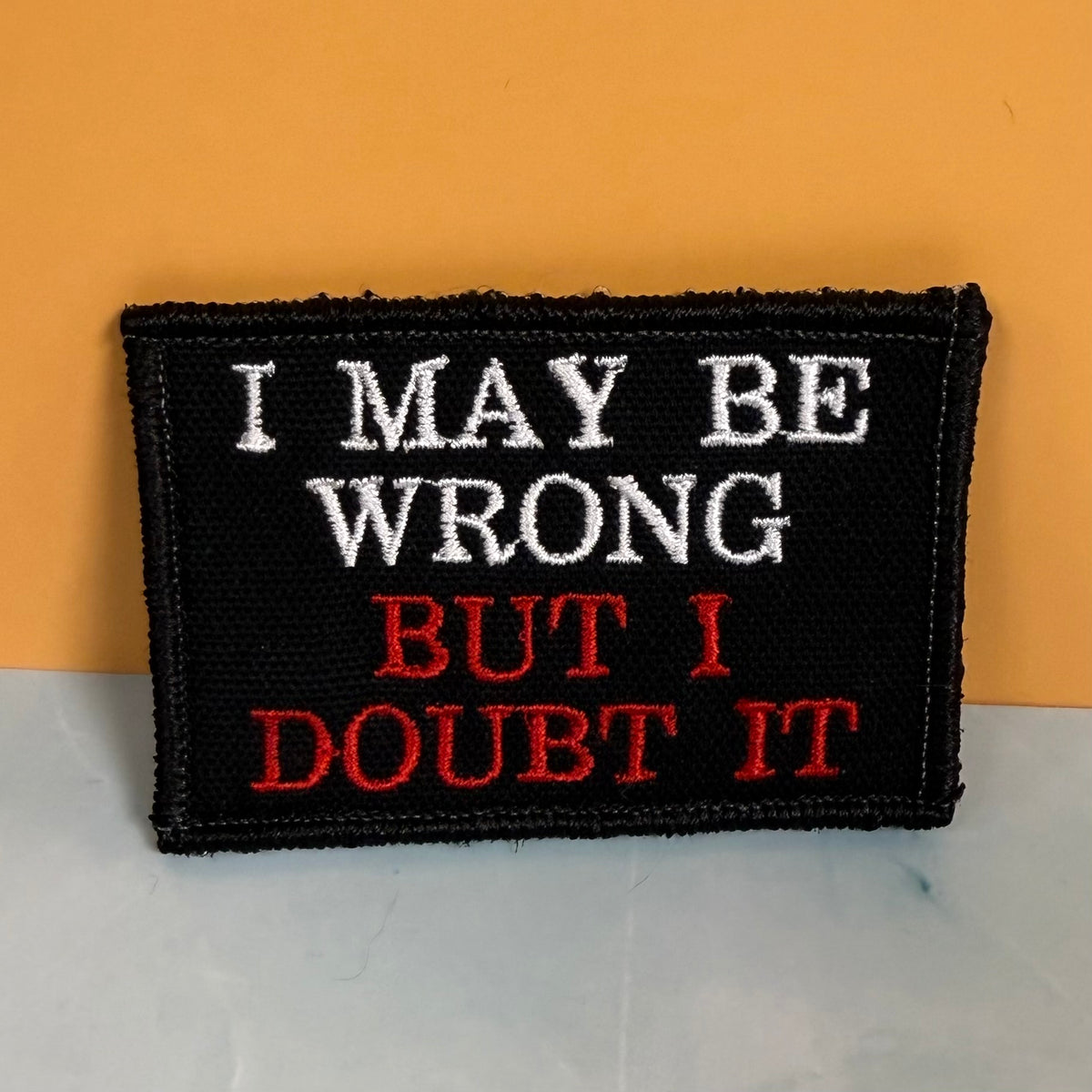 A rectangular black embroidered patch with the phrase 'I MAY BE WRONG BUT I DOUBT IT' in white and red lettering, photographed against a two-tone orange and light gray background.