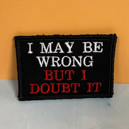 A rectangular black embroidered patch with the phrase 'I MAY BE WRONG BUT I DOUBT IT' in white and red lettering, photographed against a two-tone orange and light gray background.