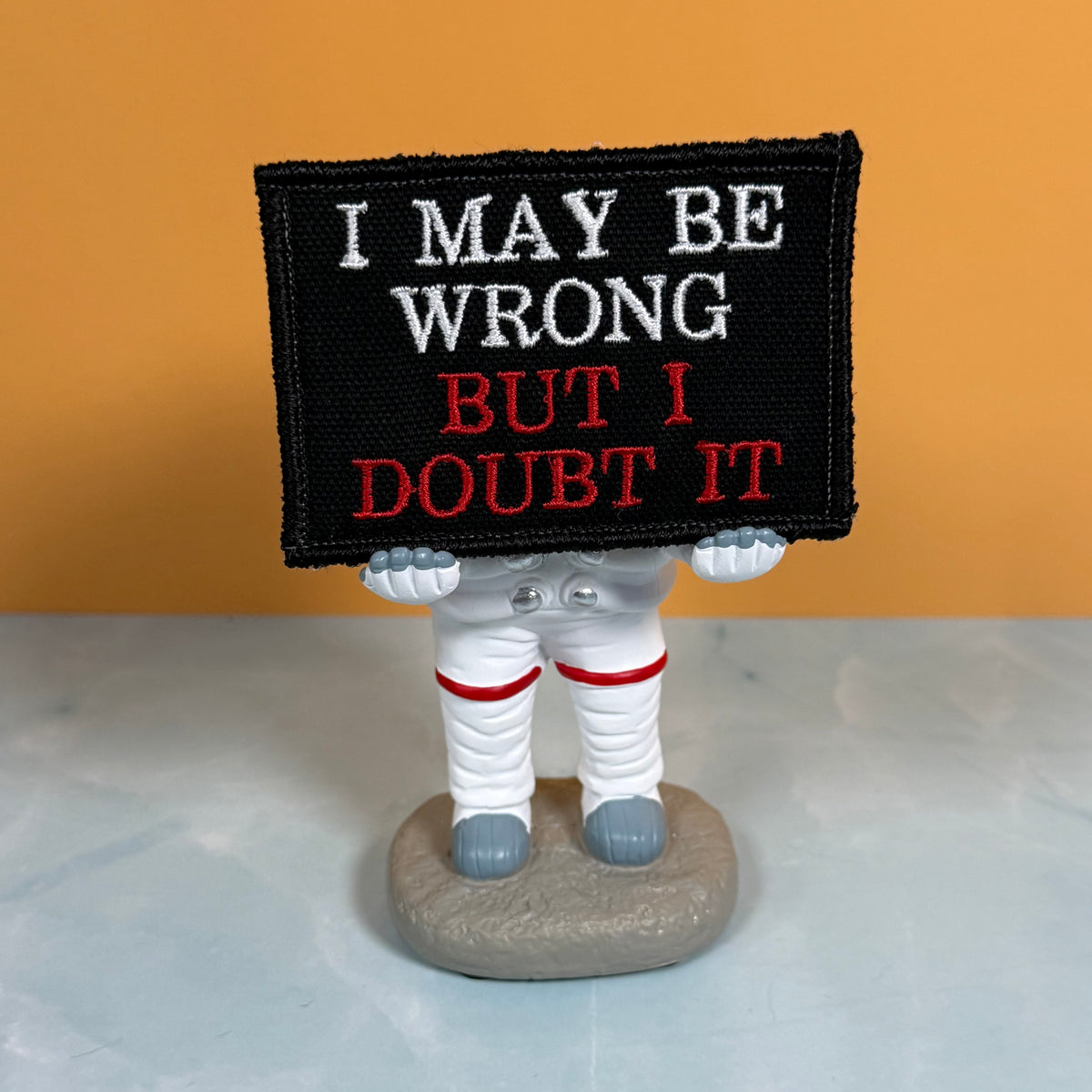 An embroidered patch with the text 'I MAY BE WRONG BUT I DOUBT IT' in white and red on a black background, held by a small astronaut figurine on a gray base against an orange and gray background.