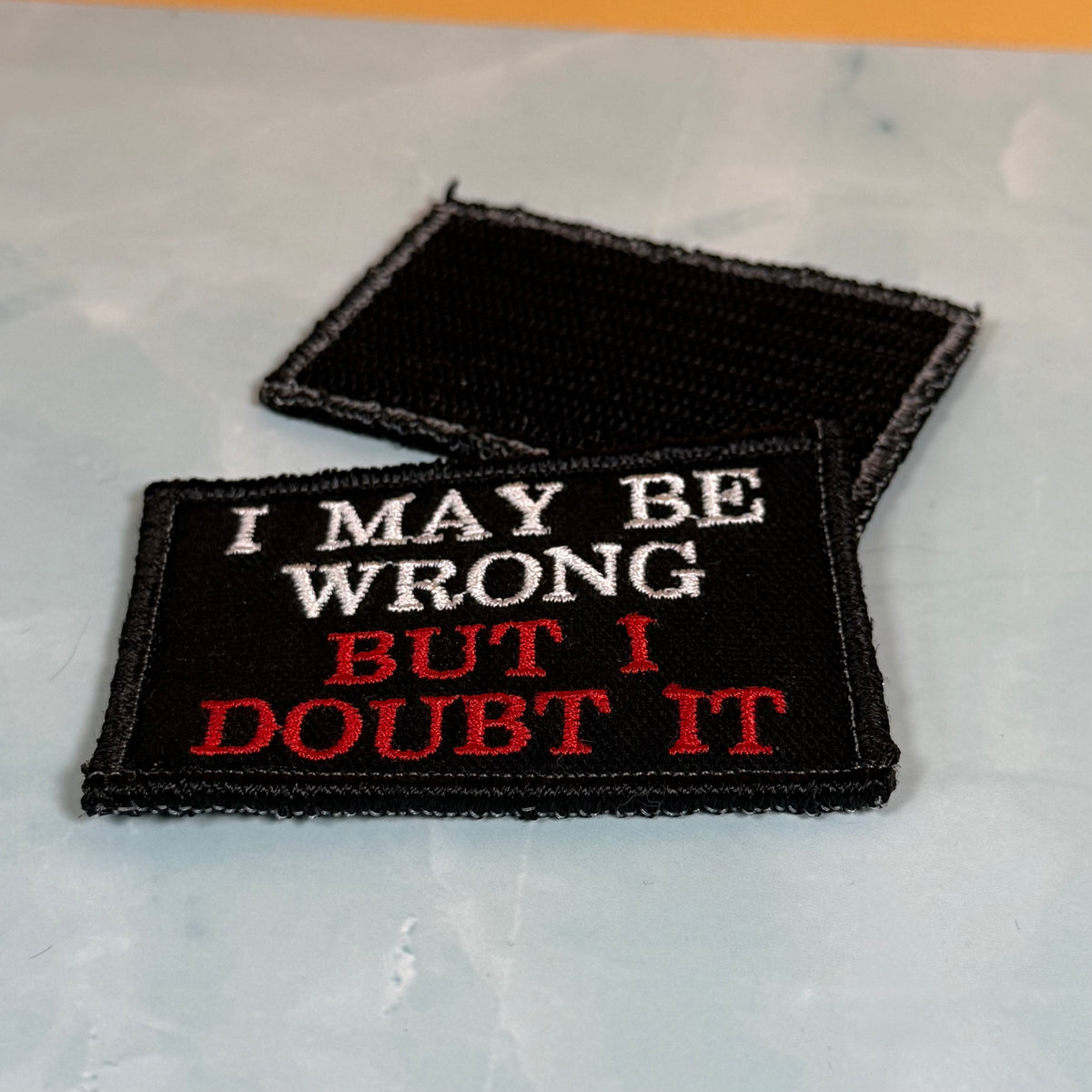 Two rectangular embroidered patches on a light gray marble-like surface. The front patch is black with white and red text reading 'I MAY BE WRONG BUT I DOUBT IT,' and the rear patch is black with no visible text.
