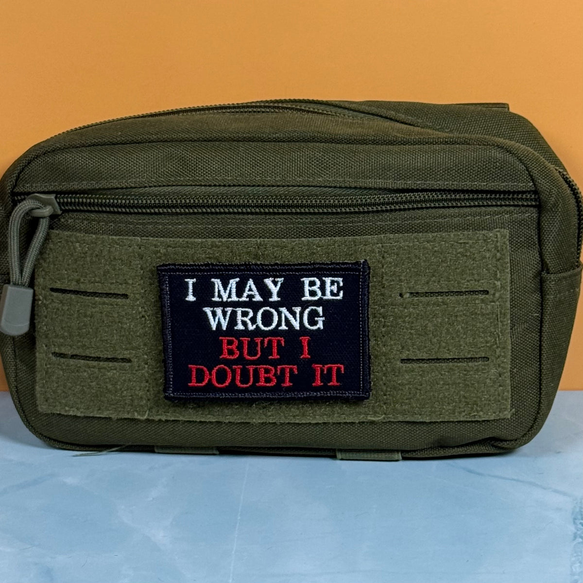 An olive green tactical pouch with a front zipper and a rectangular embroidered patch that reads 'I MAY BE WRONG BUT I DOUBT IT' in white and red text on a black background, set against an orange wall and light blue surface.