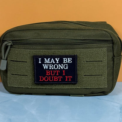 An olive green tactical pouch with a front zipper and a rectangular embroidered patch that reads 'I MAY BE WRONG BUT I DOUBT IT' in white and red text on a black background, set against an orange wall and light blue surface.