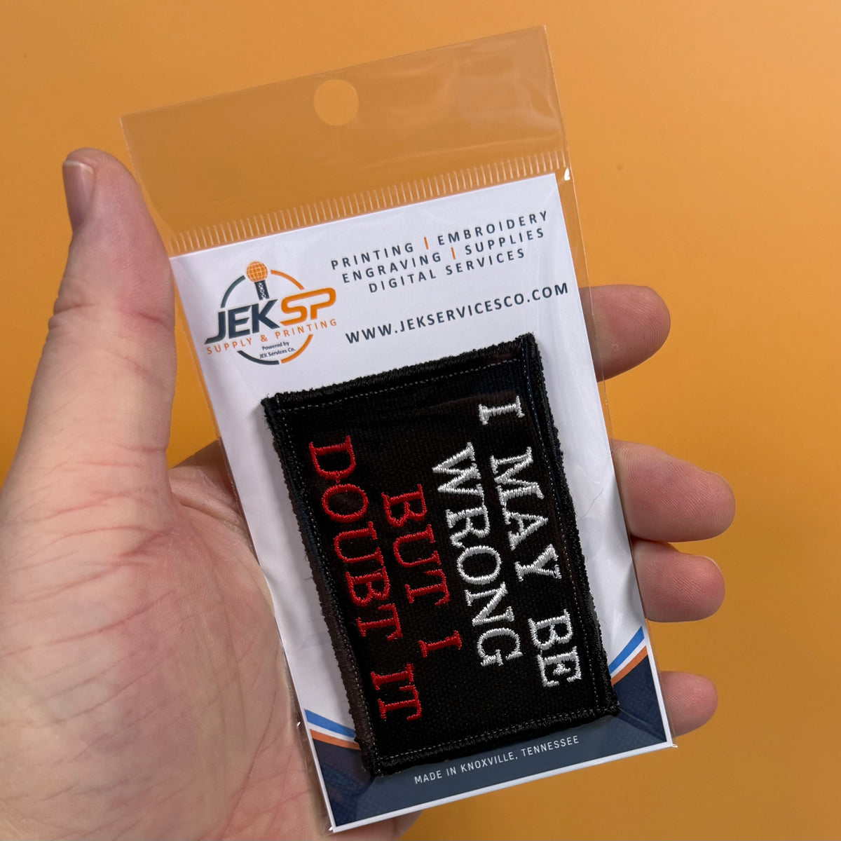 A hand holds a packaged embroidered patch against a solid orange background. The patch reads 'I MAY BE WRONG BUT I DOUBT IT' in red, white, and black text on a black square.