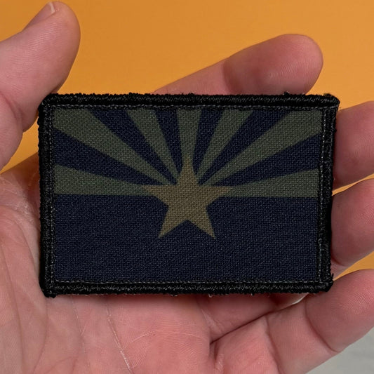 A rectangular tactical patch featuring the Arizona state flag design in dark navy blue and olive drab green, held in a person's hand against a solid orange background.