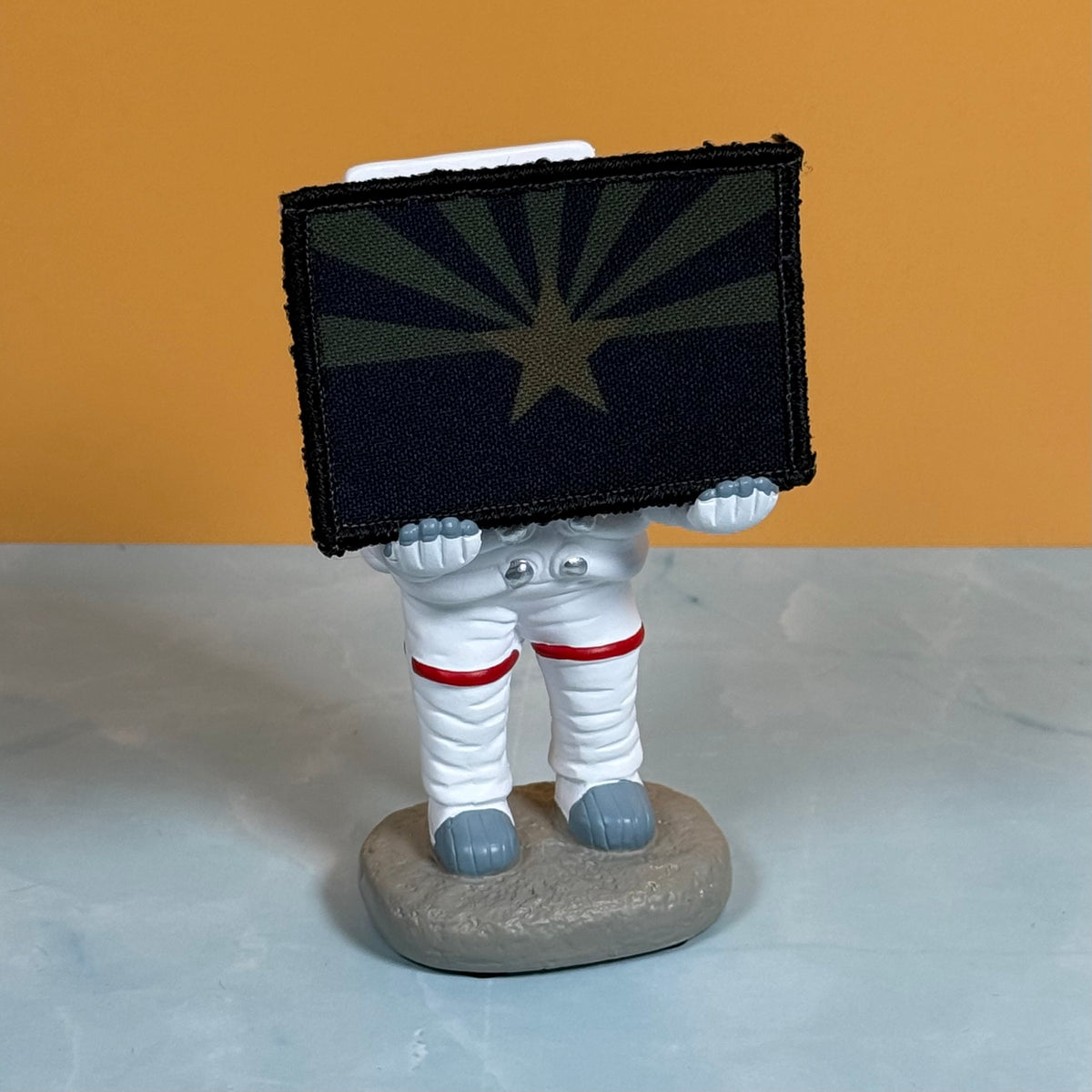 Arizona State Flag printed canvas morale patch, an astronaut figurine holding a rectangular patch with a dark navy and olive starburst design, standing on a gray base against a two-tone orange and light gray background.