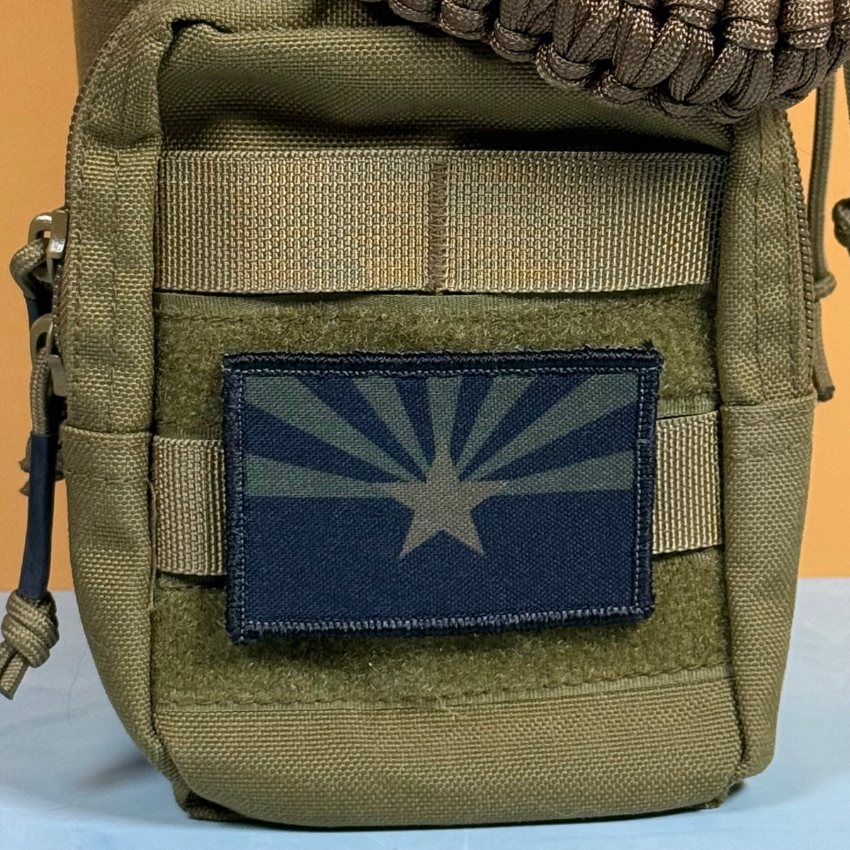 Arizona State Flag printed canvas morale patch, an olive drab tactical pouch featuring a rectangular patch of the Arizona state flag, with a paracord strap visible at the top against a solid orange background.