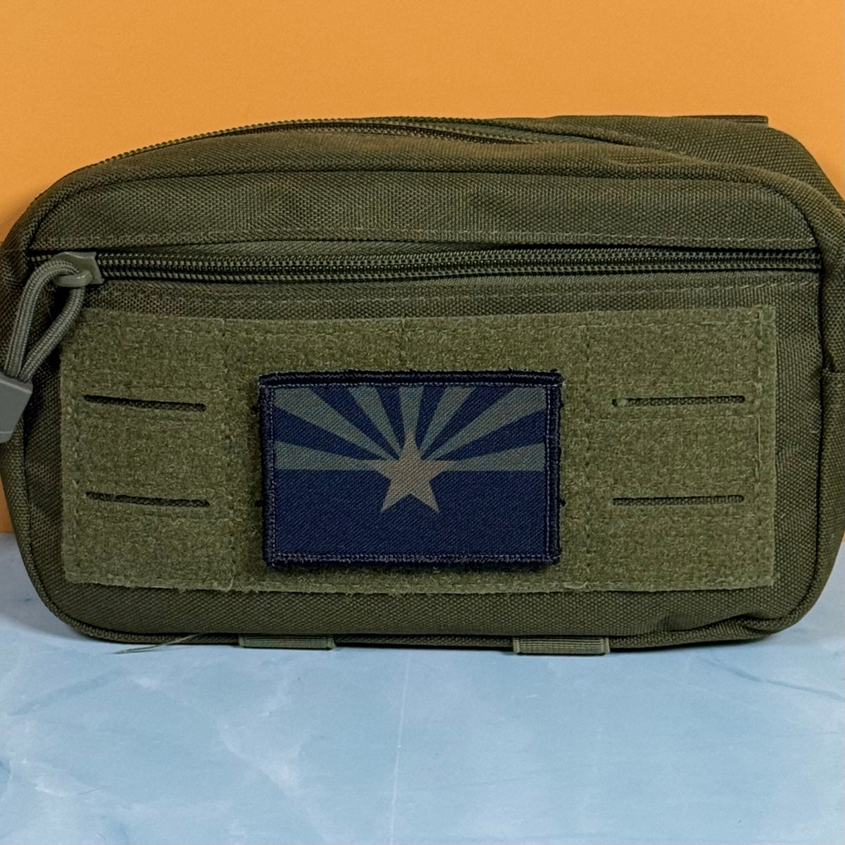 Arizona State Flag printed canvas morale patch, an olive drab tactical pouch with a front zipper and a rectangular patch featuring the Arizona state flag, set against an orange background and light blue surface.