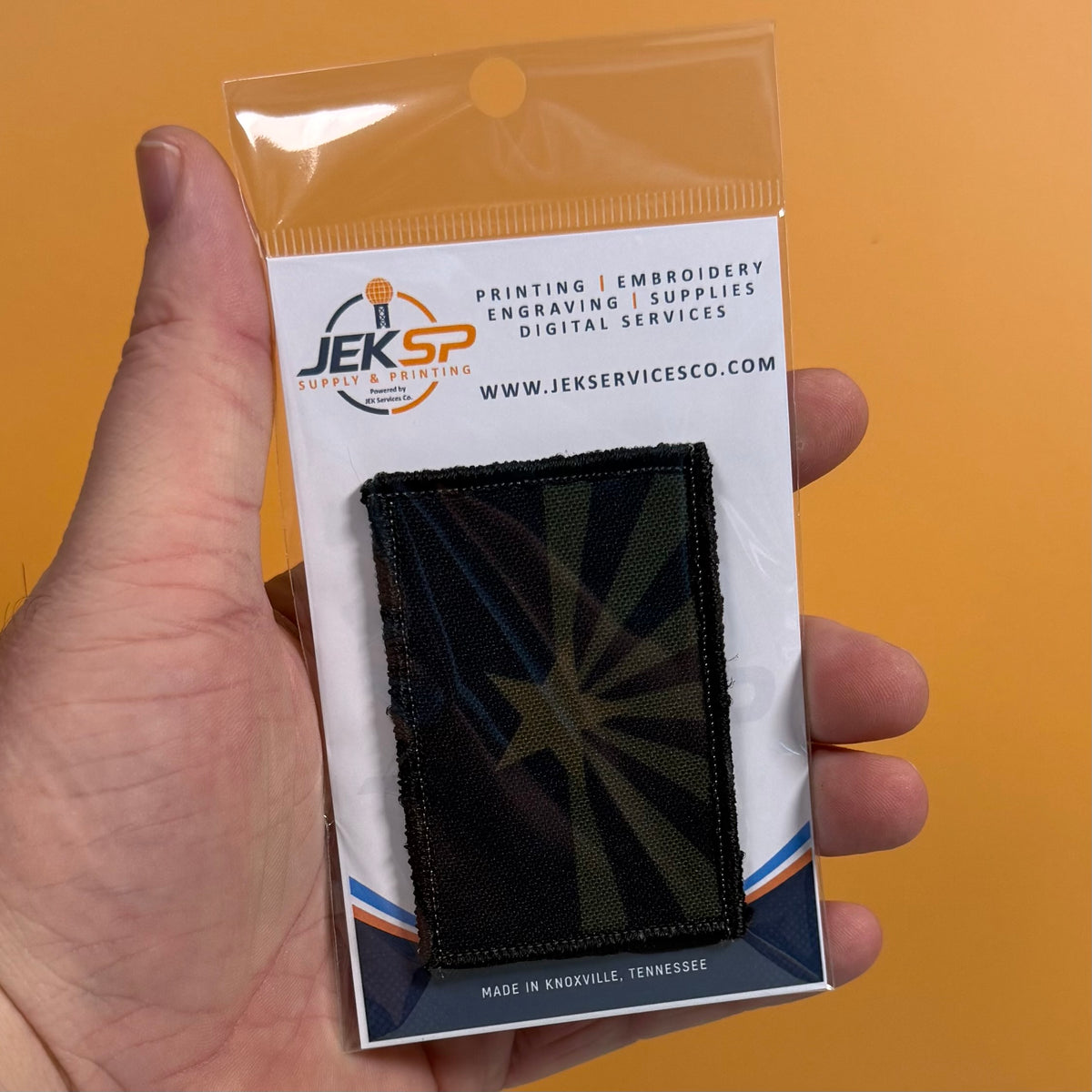 Arizona State Flag printed canvas morale patch, a hand holds a small printed patch in clear plastic packaging against a solid orange background. The patch is black with a dark green and black starburst design.