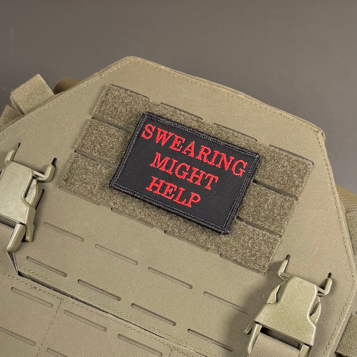 A rectangular black embroidered patch with red text reading 'SWEARING MIGHT HELP' attached to tan tactical gear with hook-and-loop panels and metal quick-release buckles.