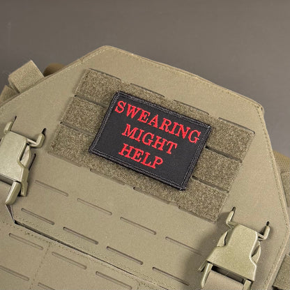A rectangular black embroidered patch with red text reading 'SWEARING MIGHT HELP' attached to tan tactical gear with hook-and-loop panels and metal quick-release buckles.