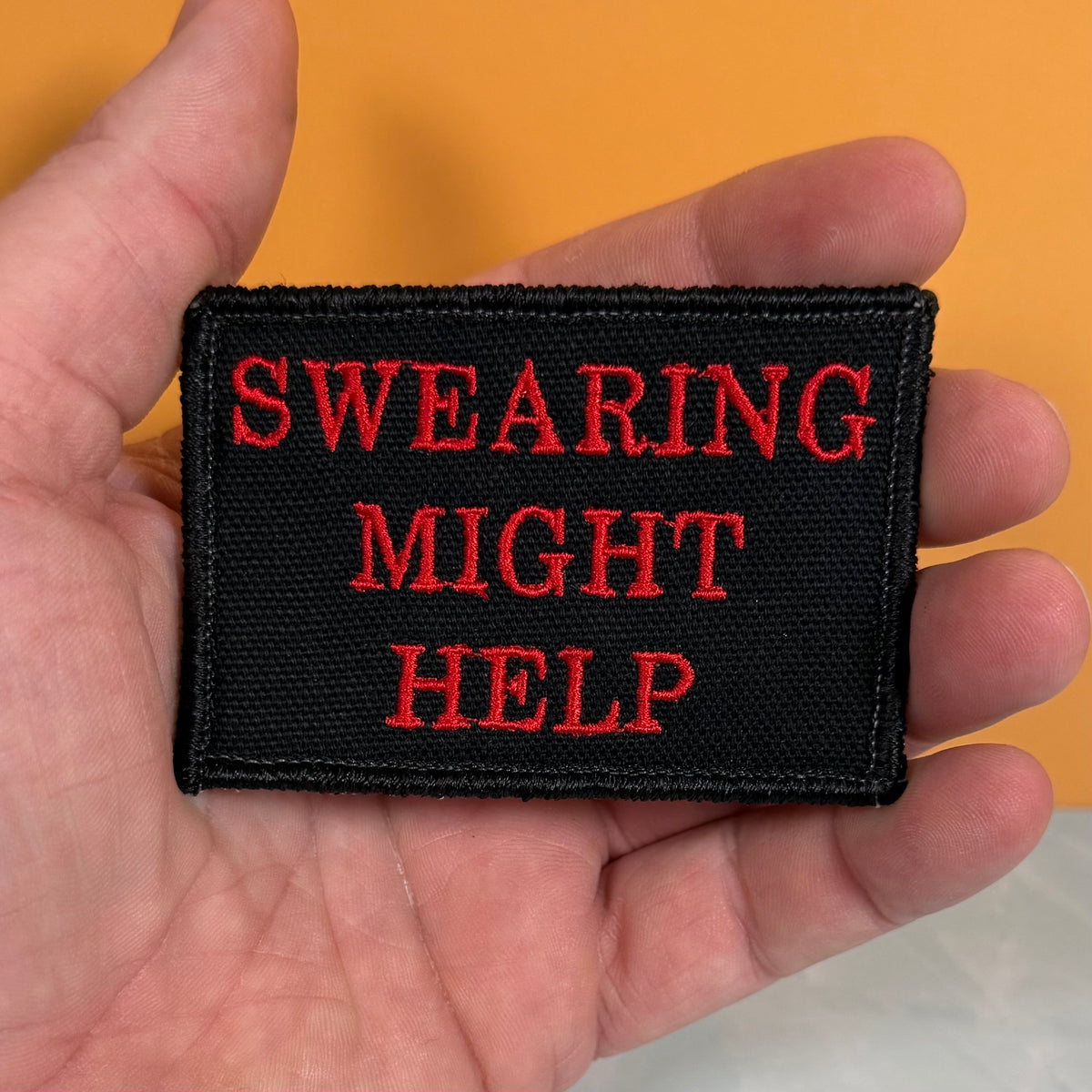 A rectangular black embroidered patch with red text reading 'SWEARING MIGHT HELP' held in a person's hand against a solid orange background.