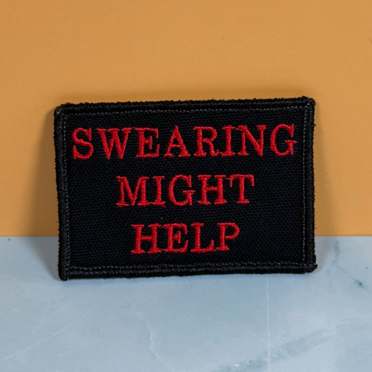 A rectangular black embroidered patch with the phrase 'SWEARING MIGHT HELP' in red lettering, displayed against a split background of orange and light gray.