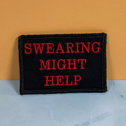A rectangular black embroidered patch with the phrase 'SWEARING MIGHT HELP' in red lettering, displayed against a split background of orange and light gray.