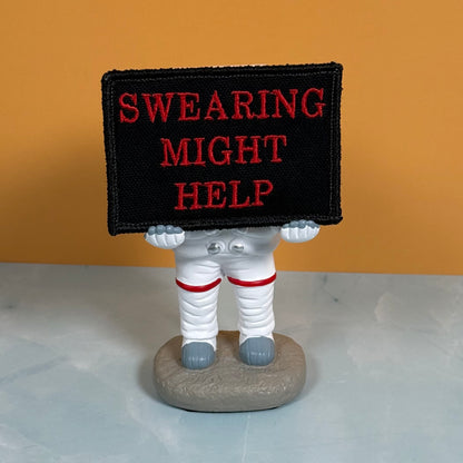 A novelty astronaut figurine holding a black rectangular sign with red embroidered text that reads 'SWEARING MIGHT HELP', set against a warm orange background and light gray surface.