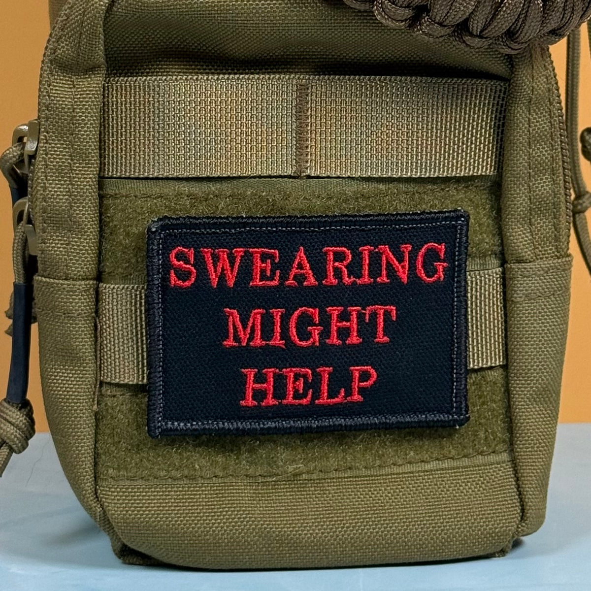 An olive green tactical pouch featuring a black rectangular patch with red embroidered text that reads 'SWEARING MIGHT HELP'.