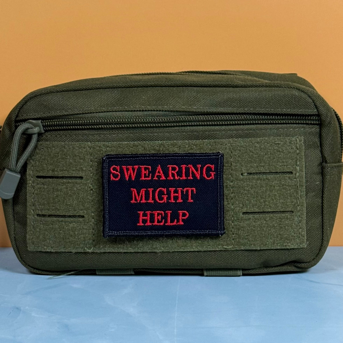 An olive green tactical pouch with a front hook-and-loop patch featuring the red embroidered text 'SWEARING MIGHT HELP' on a dark navy background.