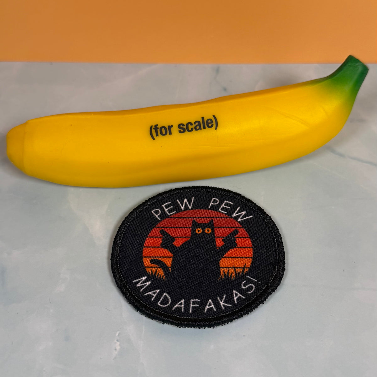 Pew Pew Madafakas Cat printed canvas morale patch, a yellow banana-shaped novelty item with green tips and the design lettering lies on a light marble-like surface against an orange background. In front of it is a round.