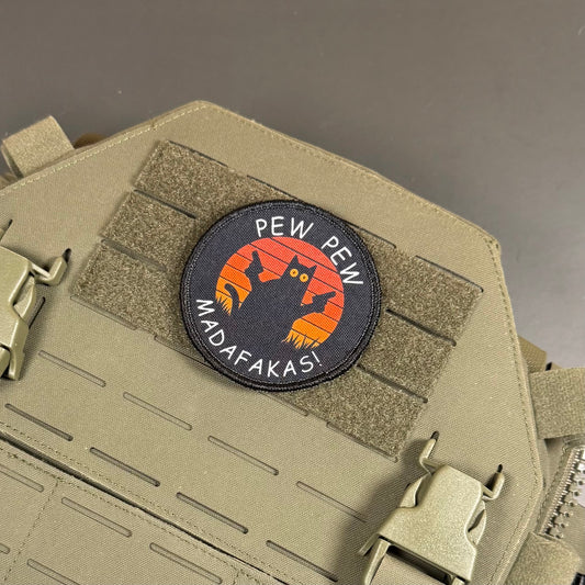 A circular embroidered tactical patch with a black cat silhouette against an orange sunset, bordered in black and reading 'PEW PEW MADAFAKASI' in white, shown attached to a tan tactical vest with MOLLE webbing.