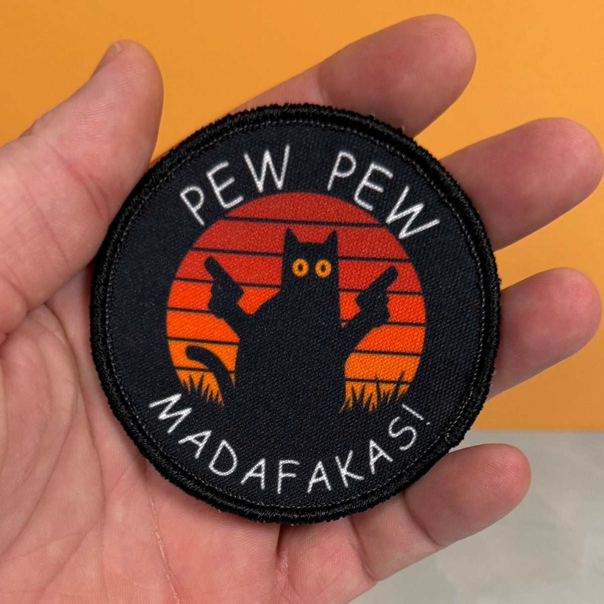 Pew Pew Madafakas Cat printed canvas morale patch held in a hand, featuring a black cat silhouette with guns against an orange sunset background and the design lettering in white.