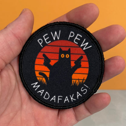 Pew Pew Madafakas Cat printed canvas morale patch held in a hand, featuring a black cat silhouette with guns against an orange sunset background and the design lettering in white.