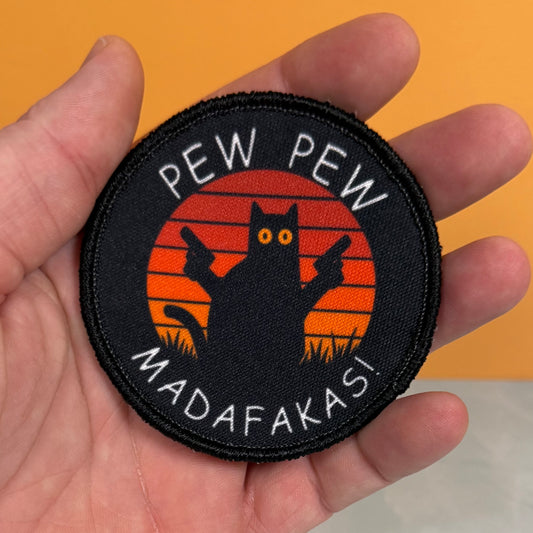 A circular embroidered patch held in a hand, featuring a black cat silhouette with guns against an orange sunset background and the text 'PEW PEW MADAFAKASI' in white.