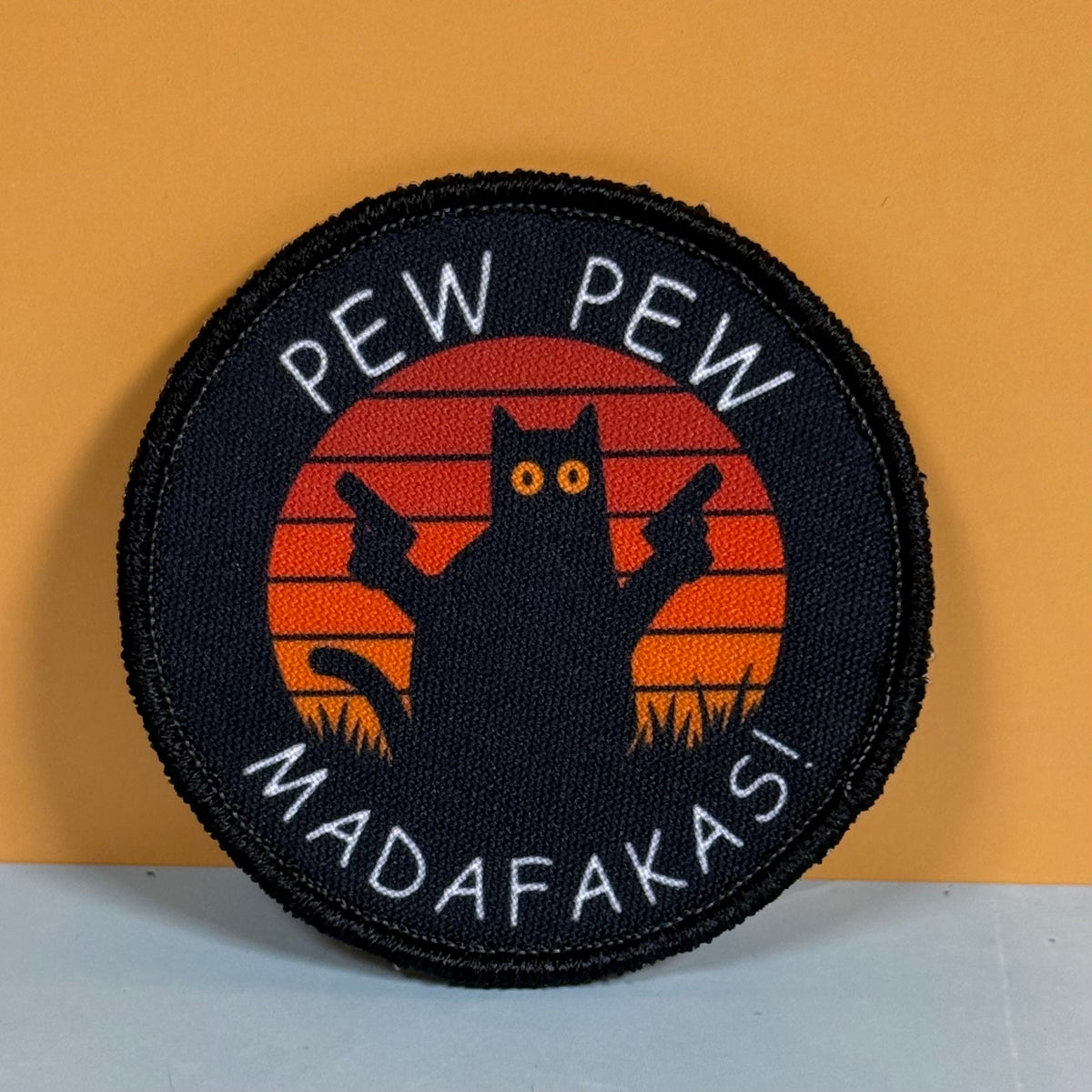 Pew Pew Madafakas Cat printed canvas morale patch, ront of an orange sunset, with the design lettering in white around the border, shown against a yellow-orange background.
