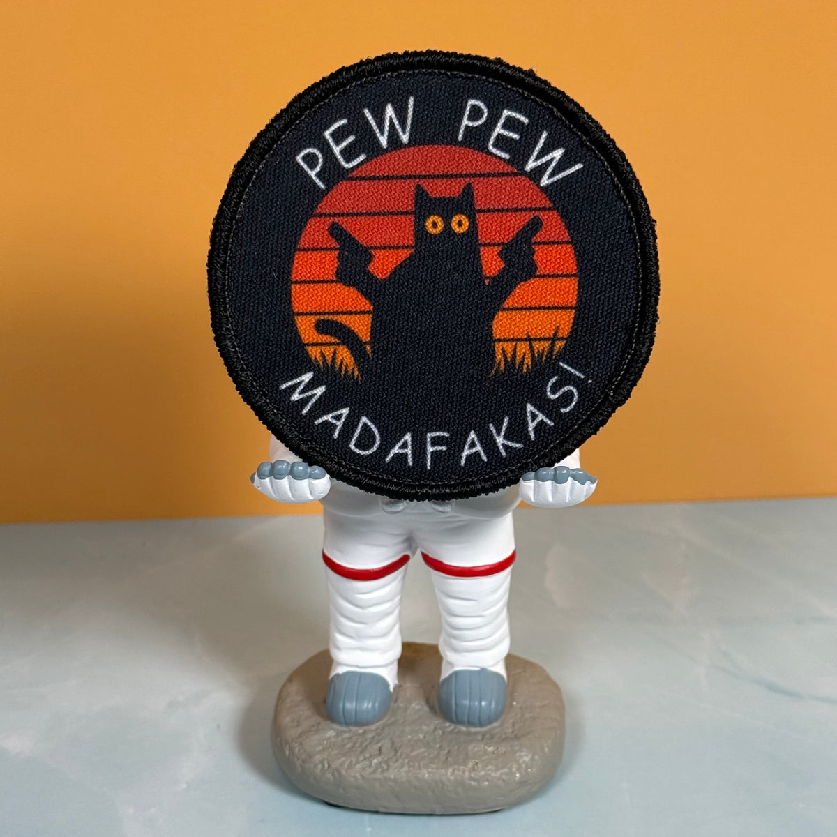 Pew Pew Madafakas Cat printed canvas morale patch, a small astronaut figurine holding a round printed patch. The patch has a black background with a black cat silhouette against orange-red sunset stripes and the design lettering.