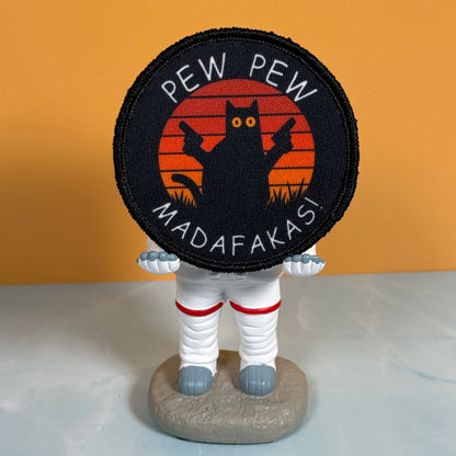 Pew Pew Madafakas Cat printed canvas morale patch, a small astronaut figurine holding a round printed patch. The patch has a black background with a black cat silhouette against orange-red sunset stripes and the design lettering.