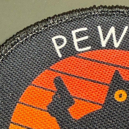 Pew Pew Madafakas Cat printed canvas morale patch, close-up of a woven fabric patch showing the letters 'PEW' in white above an orange and black graphic on a dark gray background.