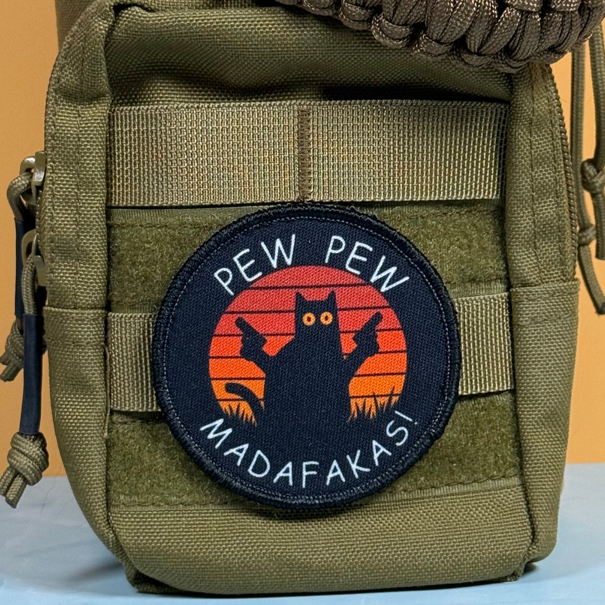 Pew Pew Madafakas Cat printed canvas morale patch, an olive drab tactical pouch featuring a circular printed patch with a black cat silhouette holding two guns against an orange sunset, with the design lettering in white.