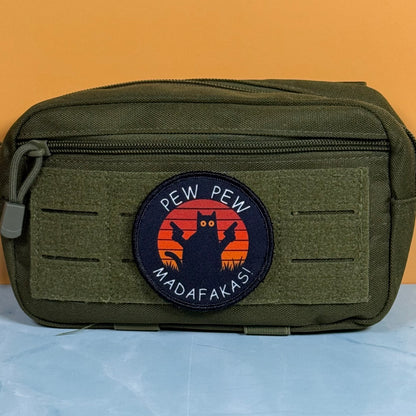 Pew Pew Madafakas Cat printed canvas morale patch, an olive green tactical pouch with a circular printed patch the design lettering featuring a black cat silhouette against an orange sunset, set against a solid orange background.