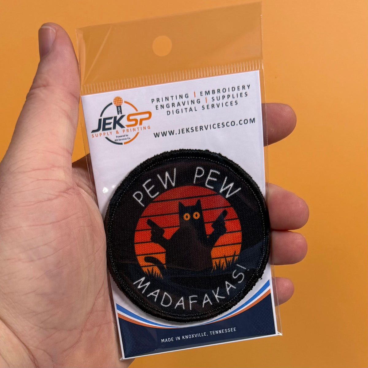 Pew Pew Madafakas Cat printed canvas morale patch, a hand holds a packaged circular printed patch against a solid orange background. The patch the design lettering with a black cat silhouette on an orange sunset-style circle.