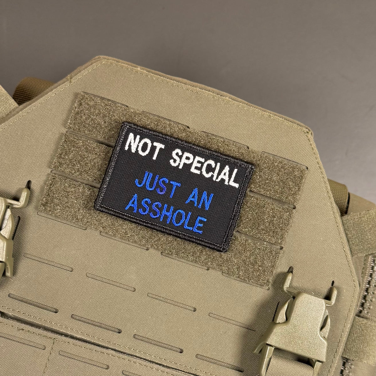 A rectangular embroidered patch with the text 'NOT SPECIAL JUST AN ASSHOLE' in white and blue on a black background, attached to tan tactical gear with velcro and quick-release buckles.