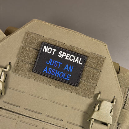 A rectangular embroidered patch with the text 'NOT SPECIAL JUST AN ASSHOLE' in white and blue on a black background, attached to tan tactical gear with velcro and quick-release buckles.