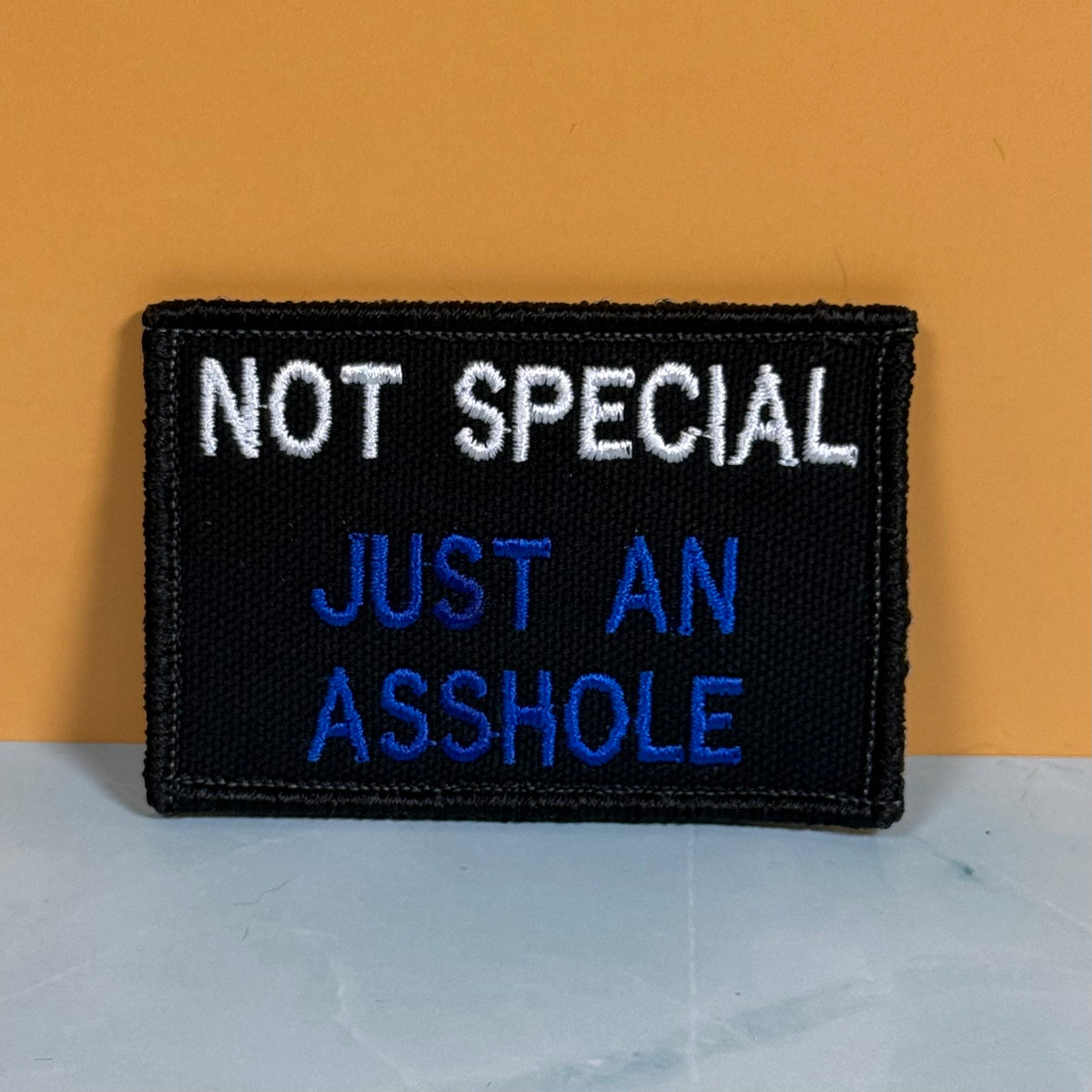 A rectangular embroidered patch with a black background and white and blue text reading 'NOT SPECIAL JUST AN ASSHOLE', photographed against a warm orange wall and pale gray surface.