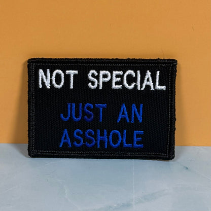 A rectangular embroidered patch with a black background and white and blue text reading 'NOT SPECIAL JUST AN ASSHOLE', photographed against a warm orange wall and pale gray surface.
