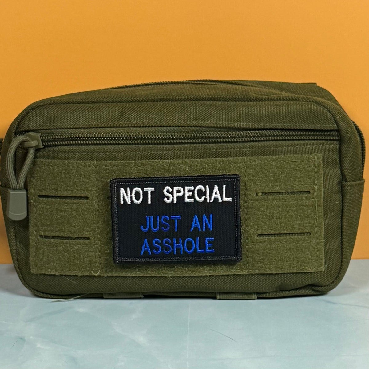 An olive green tactical pouch with a front zipper and a black embroidered patch that reads 'NOT SPECIAL JUST AN ASSHOLE' in white and blue text, set against a solid orange background.