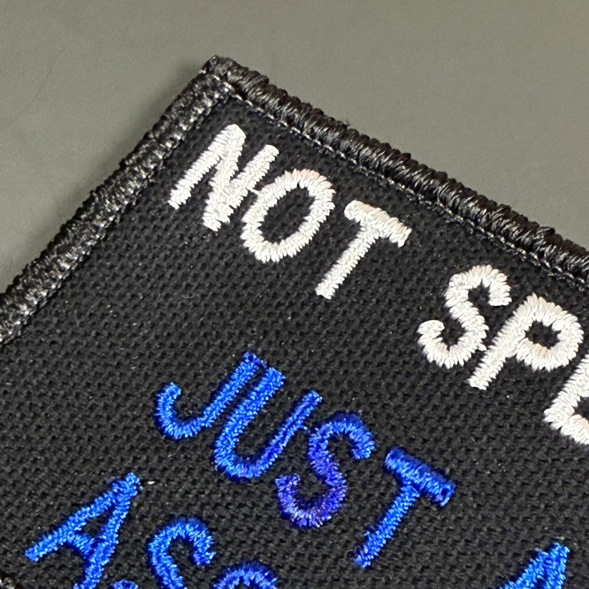 Close-up of a black embroidered patch with white and blue text reading 'NOT SPEAK JUST AS A MUTE' on a gray background.