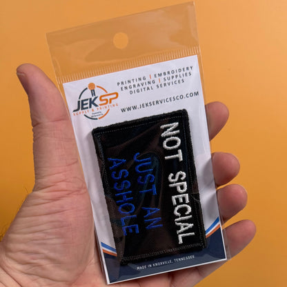 A hand holding a clear plastic retail package containing a black embroidered patch with white and blue text reading 'NOT SPECIAL JUST AN ASSHOLE.' The package card shows the JEKSP brand and service offerings, against a solid orange background.