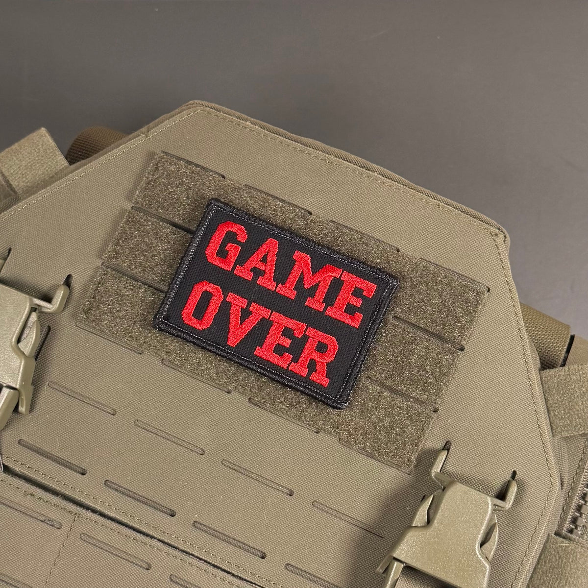 A tactical patch with the words 'GAME OVER' in red embroidered letters on a black background, attached to a tan coyote brown MOLLE vest or plate carrier.