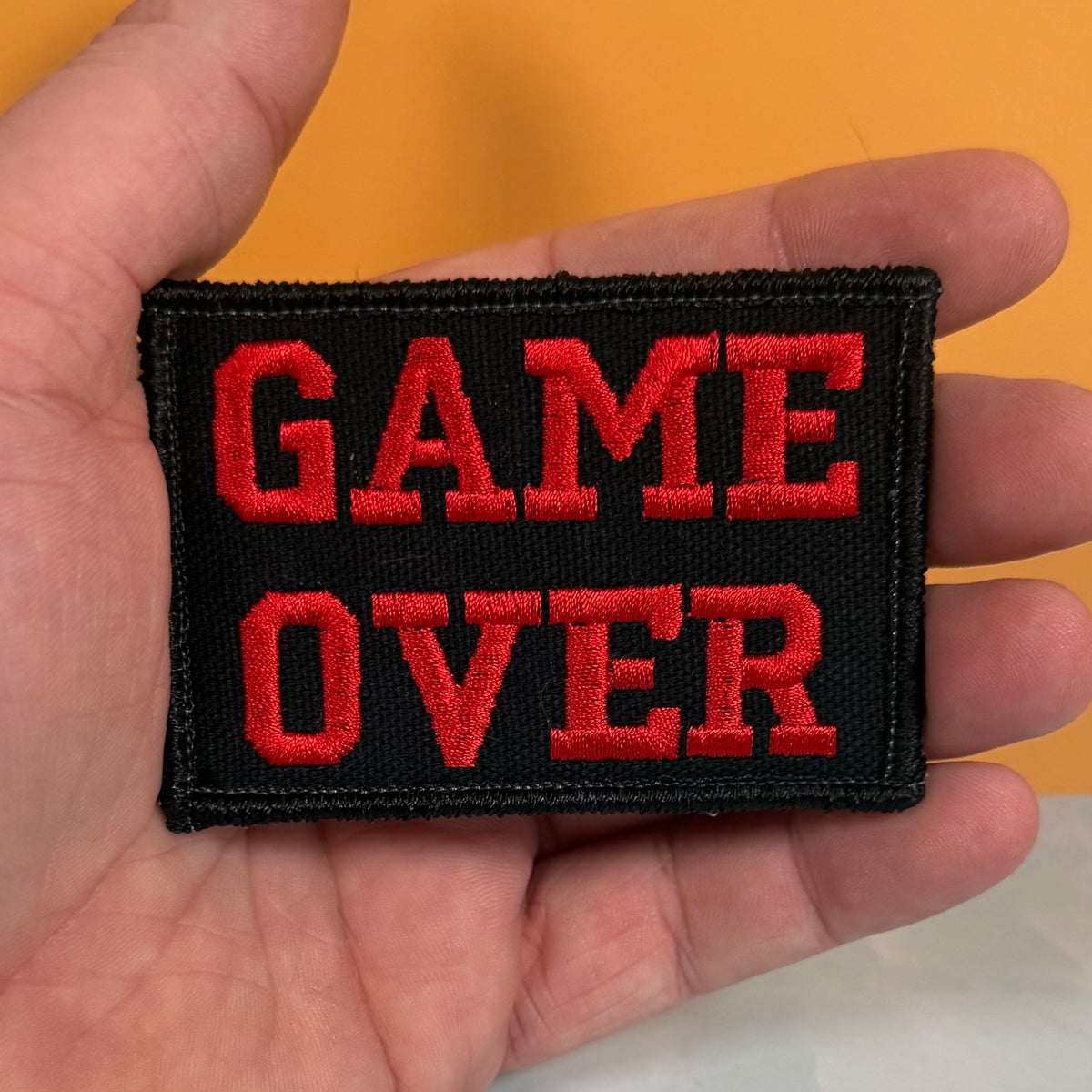 A rectangular black embroidered patch with the words 'GAME OVER' in bold red letters, held in a person's hand against a solid orange background.