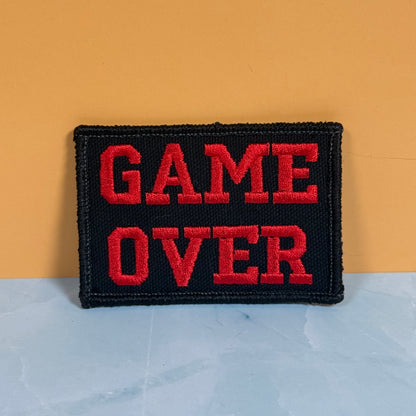 A rectangular black embroidered patch with the words 'GAME OVER' in bold red letters, standing upright against a two-tone background of mustard yellow and pale blue.