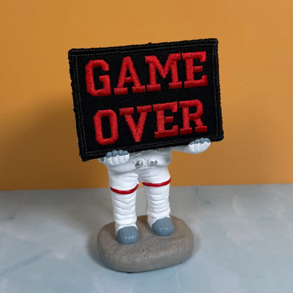A small astronaut figurine holding a black rectangular sign with red embroidered text reading “GAME OVER,” set against a warm orange background and light gray surface.